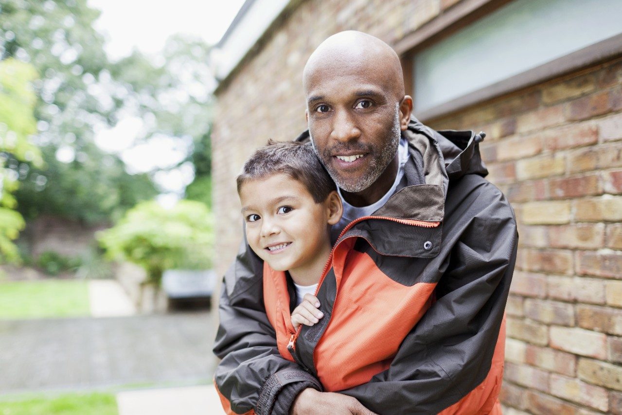 Adult man protecting boy with his warm rain jacket in front of brick-built façade