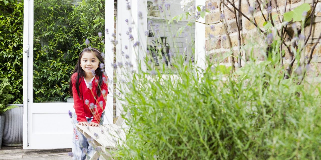 Smiling girl standing on patio leaning against a wooden bench in front of brick-lined wall